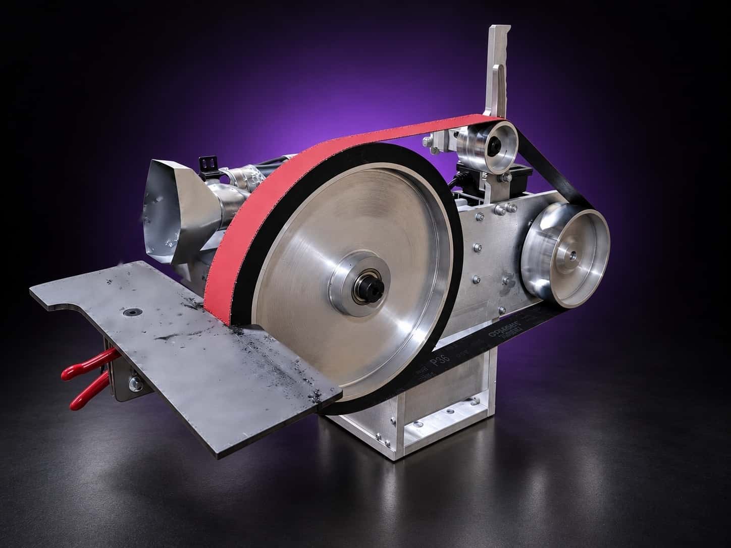 Large aluminum contact wheel with black rubber rim and red belt, steel work table in front, on black with a purple glow